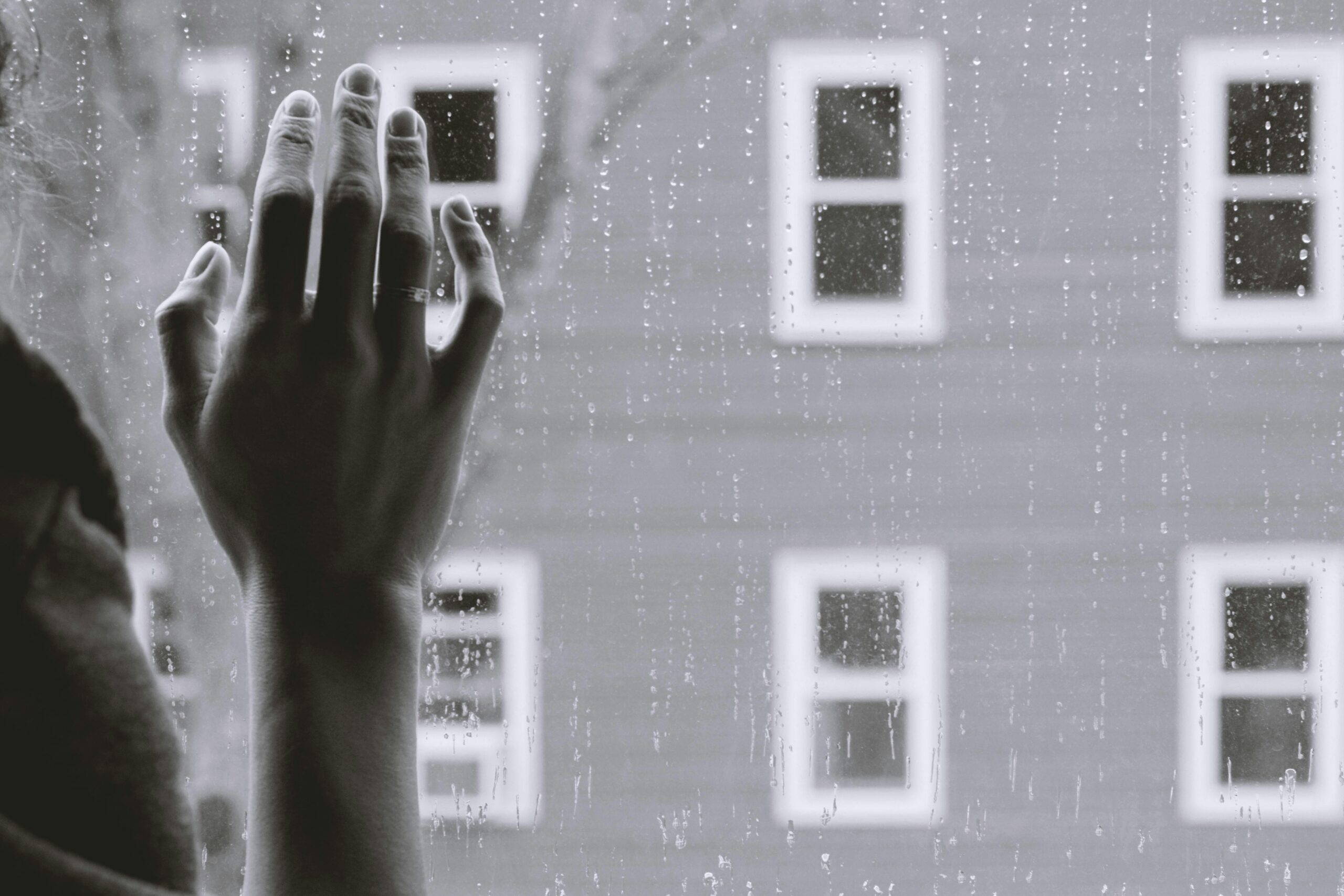 Black and white photo of woman looking out window at a raining day.